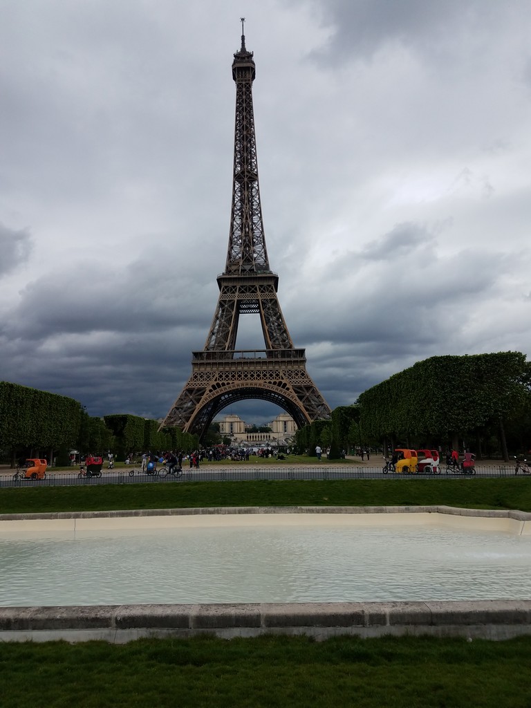 a large metal tower with a pool of water and people in the background with Eiffel Tower in the background