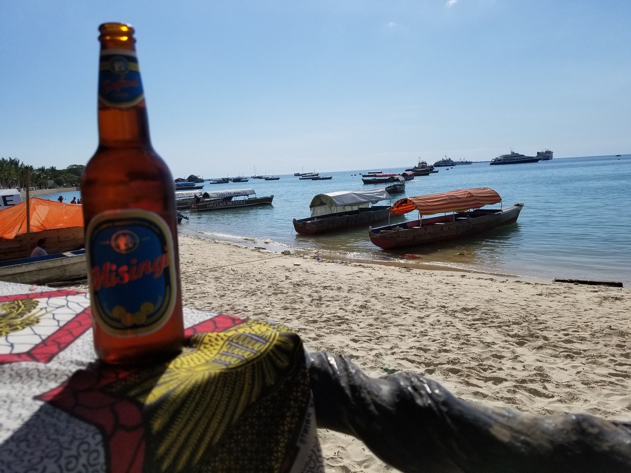 a beer bottle on a table on a beach with boats in the background