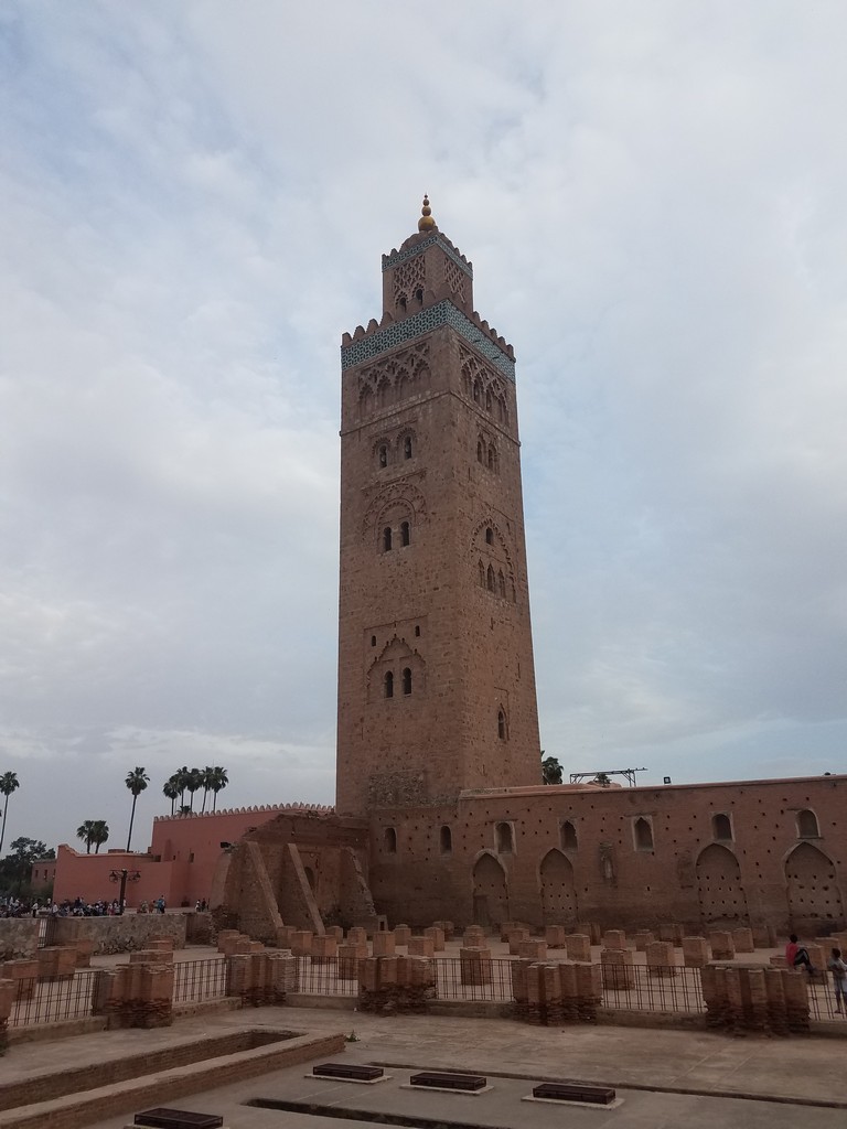 a tall tower with a wall and many people around it with Koutoubia Mosque in the background