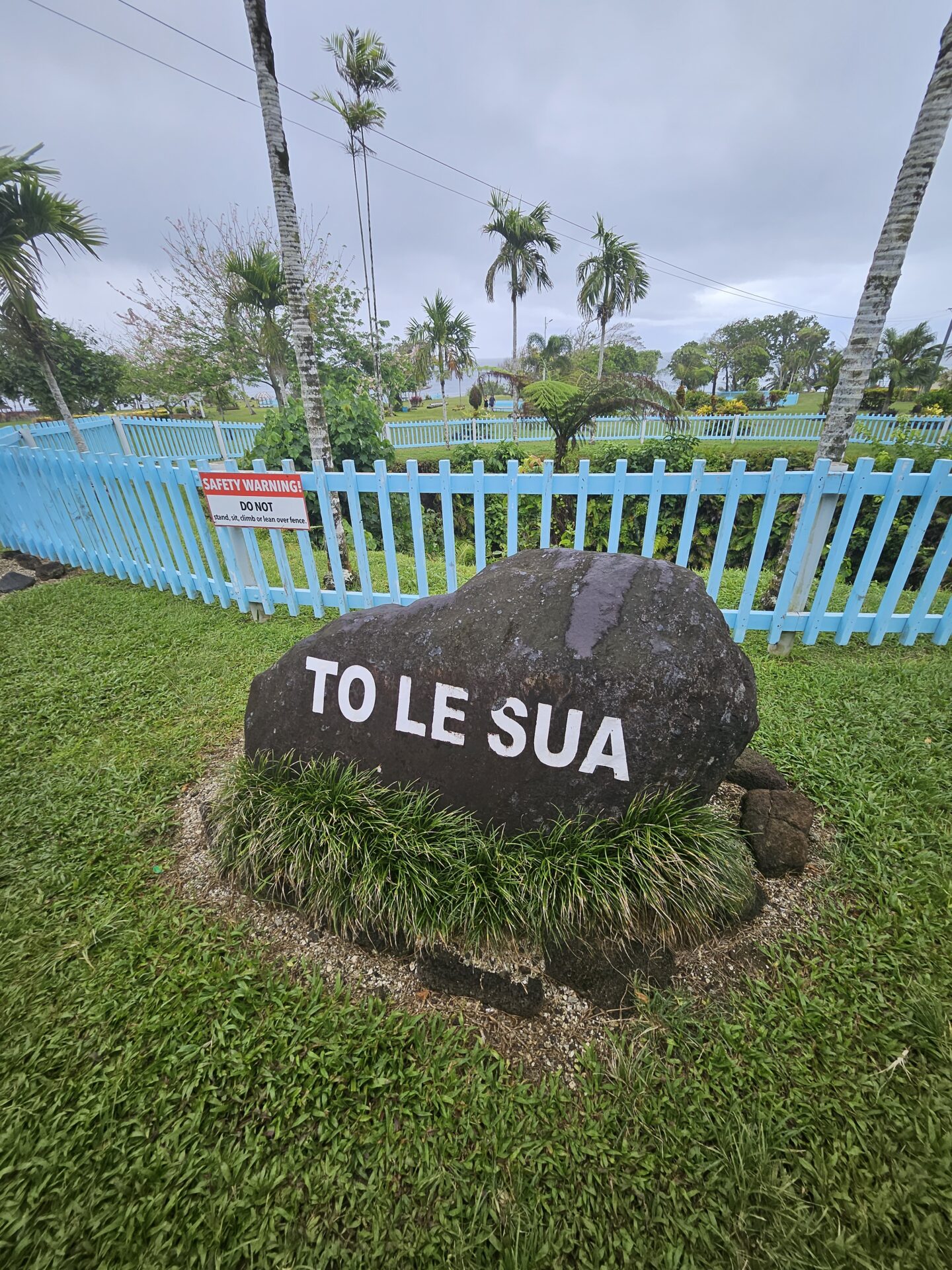 a rock with white text on it in a grassy area with a blue fence