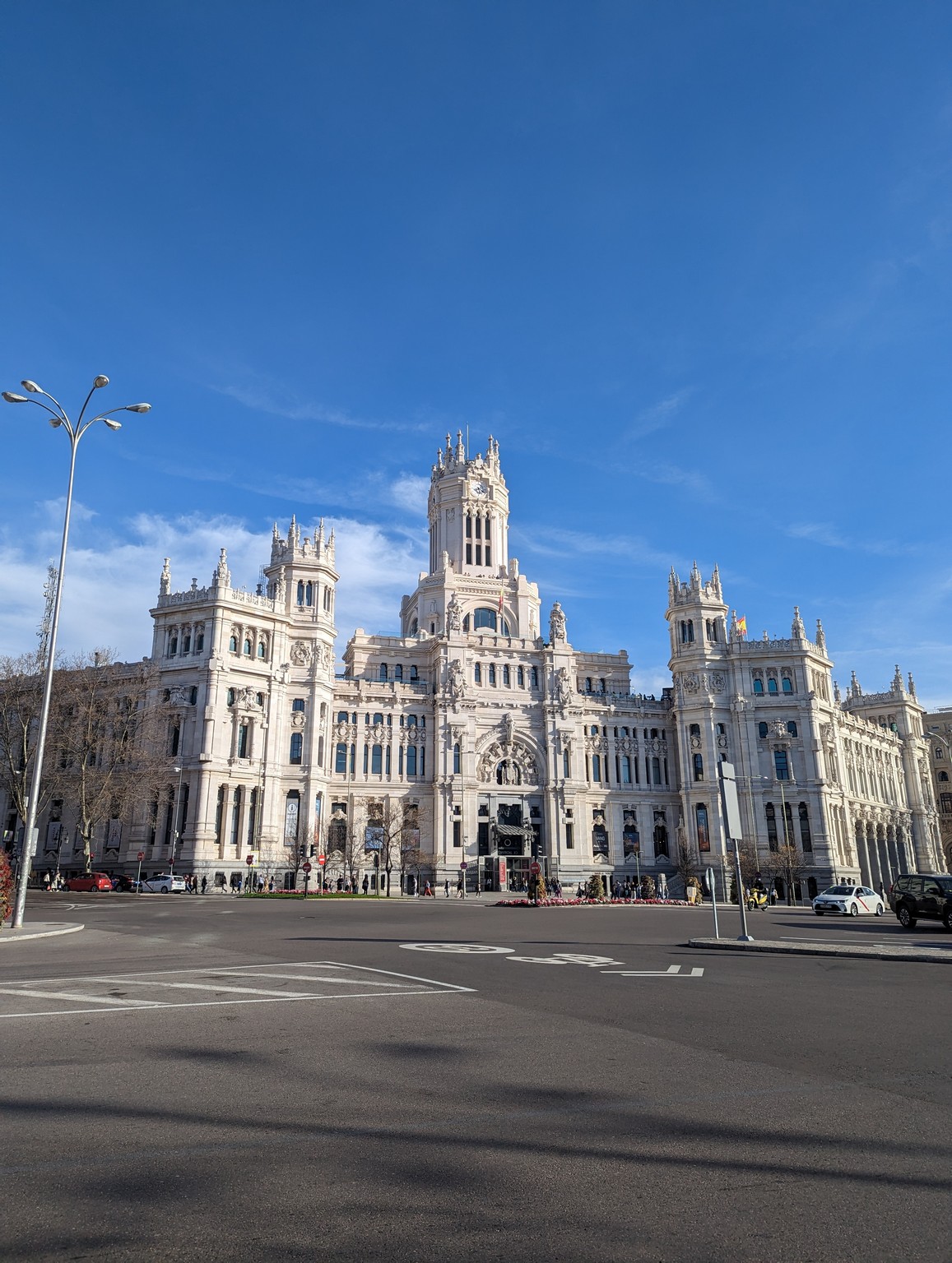 a large white building with towers and a street and a blue sky with Plaza de Cibeles in the background