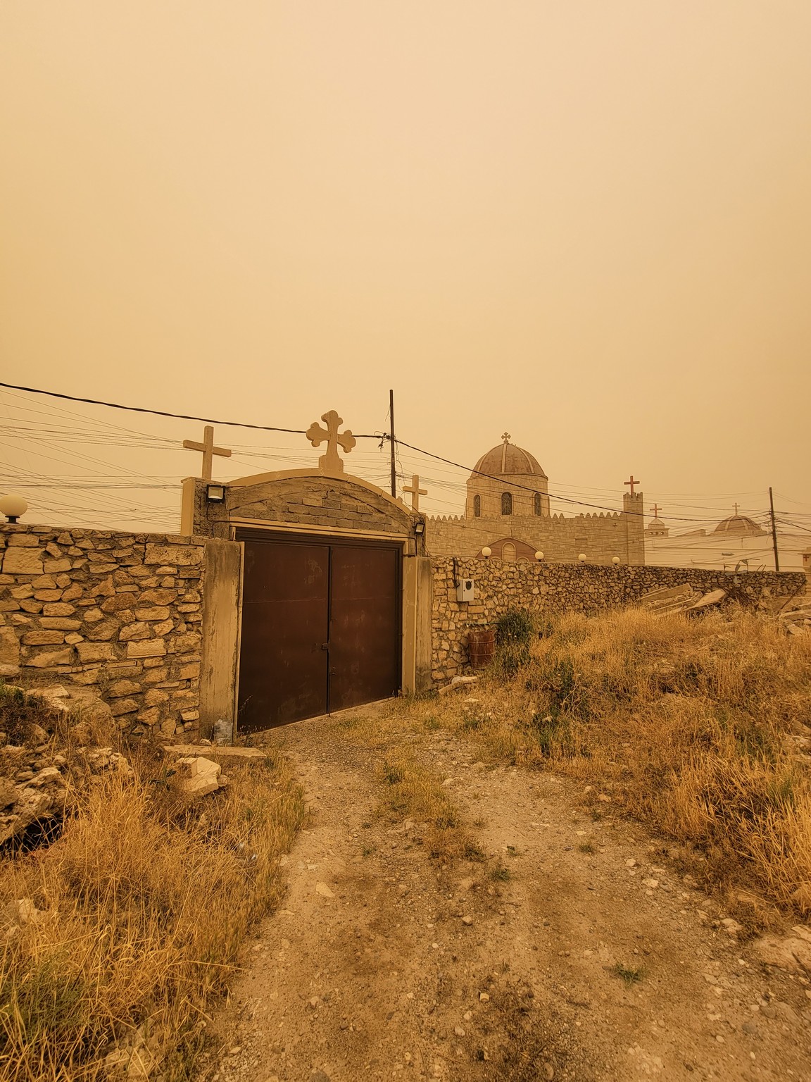 a dirt road leading to a stone wall with a gate and a building in the background